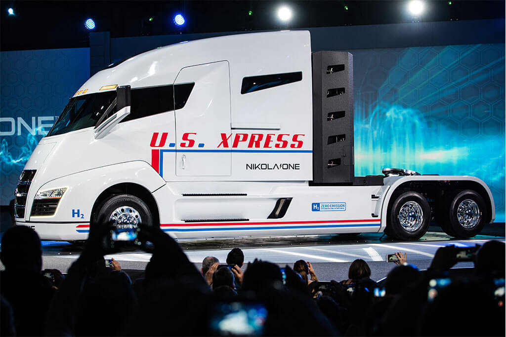 Nikola – das zweite Unternehmen am E-Mobility-Markt, das sich mit seinem Namen auf den (fast vergessenen) Elektropionier Nikola Tesla bezieht, will ganz weit hinaus: Der „Nikola One“ kombiniert Lithium-Ionen-Akkus und Wasserstoff-getriebene Brennstoffzellen. Damit soll er Reichweiten von bis zu 1.000 Meilen, also mehr als 1.600 Kilometer, erzielen. Der große Vorteil, den die Brennstoffzelle gegenüber Akkus haben: Die Tanks sind in 20 Minuten aufgefüllt. 2020 sollen die ersten Trucks ausgeliefert werden (Quelle: Nikola Motor Company). Weißer Elektro Truck Nikola One mit rotem Schriftzug in Messehalle