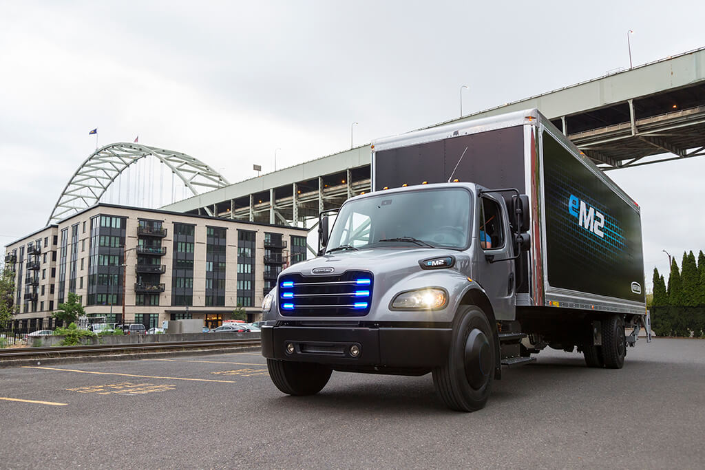 Kurz vor Weihnachten 2018 hat der Mercedes-Mutterkonzern Daimler einem Kunden in Kalifornien den ersten E-Truck der US-Marke Freightliner ausgeliefert. Insgesamt sind nach Angaben von Mercedes 20 normale LKW und zehn Sattelschlepper bestellt. Mit dieser Produktpalette nennt sich Daimler „weltweit erster Hersteller, der elektrische LKW in allen Gewichtsklassen gemeinsam mit Kunden in der Praxis erprobt“. Bei 18- bis 26-Tonnern und bei den Sattelschleppern macht den Schwaben unter anderem die VW-Tochter MAN Konkurrenz (Quelle: Daimler AG). E-Truck mit blendenden Lichtern vor einer Brücke